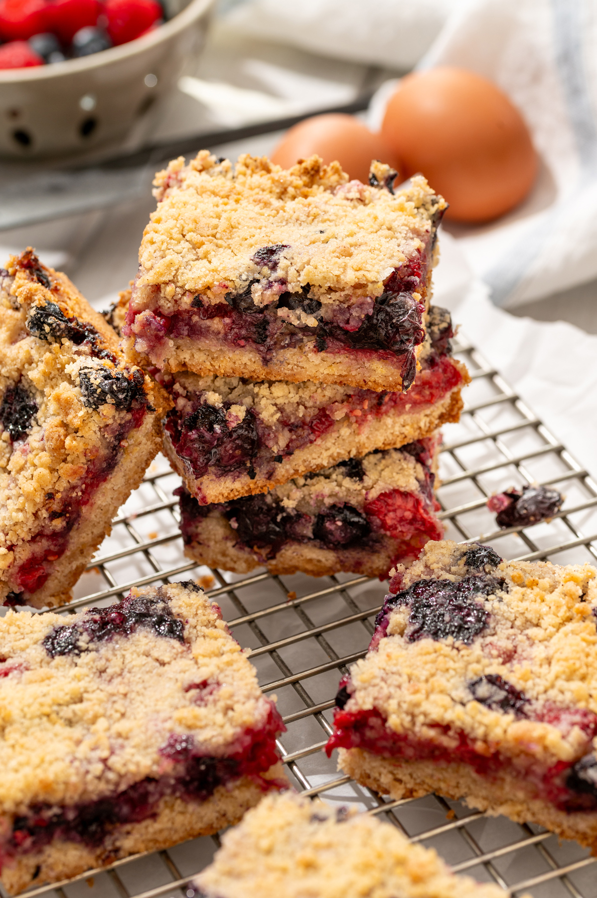 Stack of homemade berry crumble bars on cooling rack with fresh fruit and eggs in background, photographed for the fine fettle